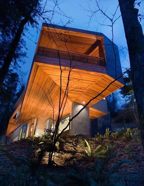 Modern wooden house with a cantilevered design, illuminated warmly at dusk in a forest setting.
